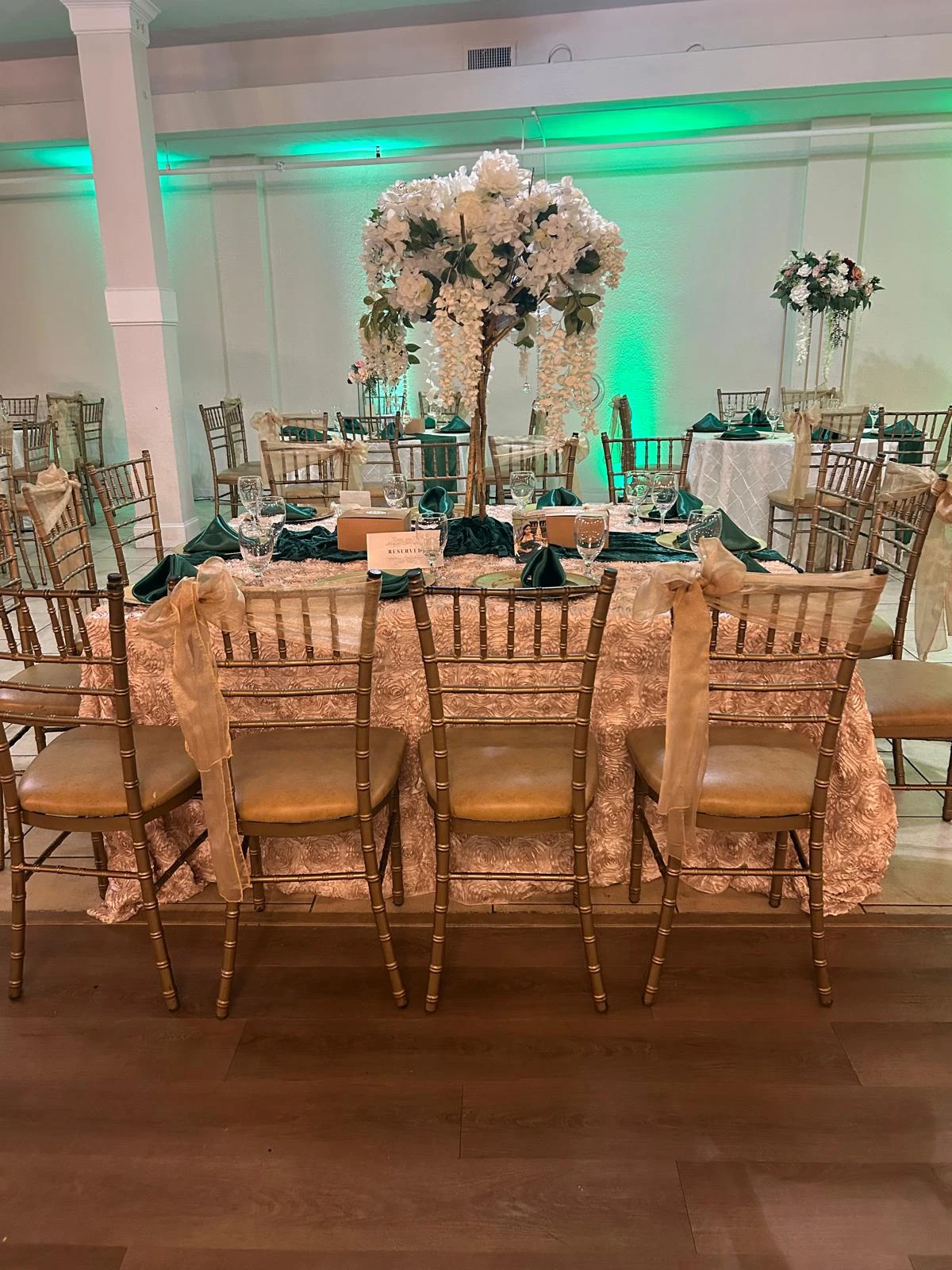 Gold table linens, Chiavari chairs and a white floral centerpiece with emerald uplighting