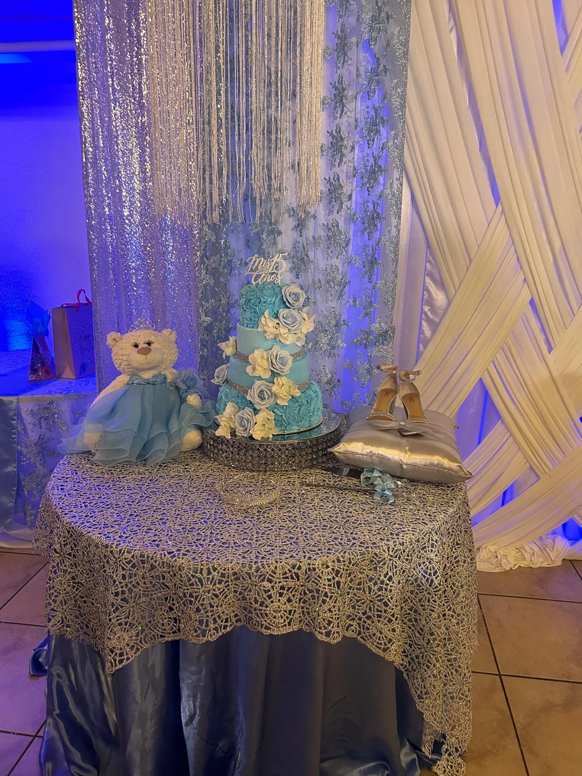 Blue and white quinceañera cake, teddy bear and shoes on a decorated cake display table